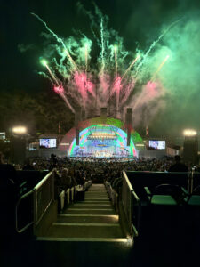 Outdoor stadium concert at night with colorful fireworks over a neon-lit stage and large crowd below.