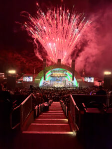 Nighttime outdoor concert with a rainbow-lit stage and a fireworks display above the crowd.