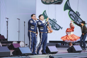 Two mariachi-style singers in blue embroidered suits sing into mics on stage, with a violinist nearby and a dancer in a bright orange dress twirling in the background.
