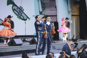 Mariachi musicians sing on stage while two violinists in blue suits perform, with colorful dancers in background.