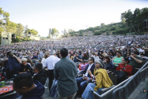 Hundreds of people seated in an outdoor amphitheater, with green hills and trees in the background, attending an event at dusk/dawn lighting.