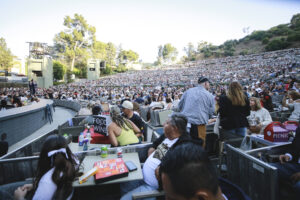 Outdoor concert venue filled with hundreds of spectators seated on tiered stands, stage left with performers.