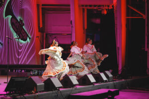 Four dancers in traditional white and orange-trimmed dresses whirl on a pink-lit stage, microphones and speakers in view.