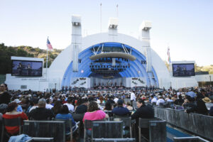 Outdoor concert at a large white amphitheater with a full band performing on a blue curved stage and a seated audience in front.