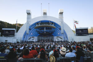 Outdoor concert at a large venue with a blue arched stage and a full audience seated in front of the performers on stage.