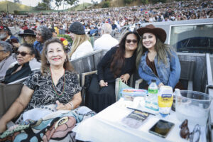 Three women in a VIP box at an outdoor concert, smiling at the camera with a packed stadium behind them and snacks on the table.