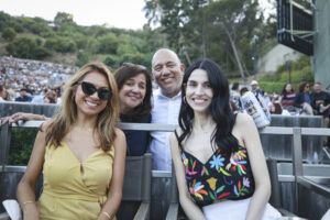 Four friends posing for a photo at an outdoor event, smiling at the camera with a crowd in the background and a beer can visible on the right side of the frame?