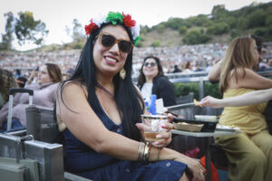 Smiling woman in a flower crown and sunglasses wearing a blue dress, holding a drink at an outdoor event with a large crowd in the background.