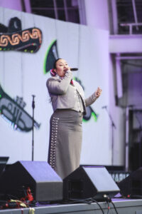 Female singer on stage wearing a gray suit, holding a microphone and singing.