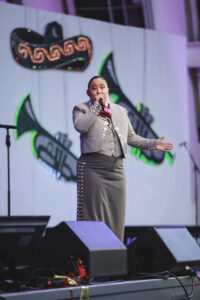 Male singer performing on stage in a gray suit with decorative trim, holding a microphone.
