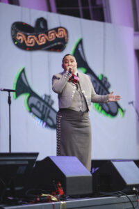 Singer in a gray suit performs on stage, singing into a microphone with a neon-backed backdrop behind him.