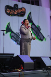 Female singer in a gray suit with pink accents performs on a stage, holding a microphone against a neon guitar backdrop.