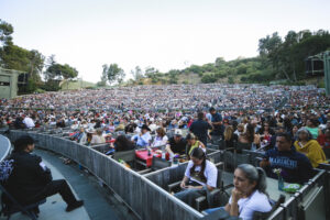 Large outdoor amphitheater crowded with hundreds of people seated in terraced rows, with trees on a hillside in the background.
