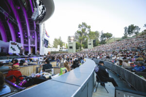 Outdoor concert at an amphitheater with a large purple-lit stage and a packed audience on tiered seating around a curved barrier.