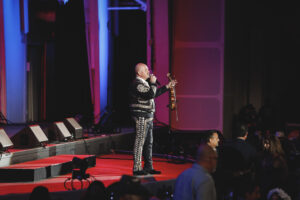 Male performer on a red-lit stage sings into a microphone, holding a violin by his side, wearing a sequined jacket with blue and purple stage lights. Audience visible in the foreground.