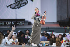 Female violinist singing into a microphone on an outdoor stage, wearing a gray dress with silver buttons; audience seated below.