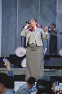 Female singer in a light gray suit performs on stage with a microphone, red bow, and pearl-embellished seams.