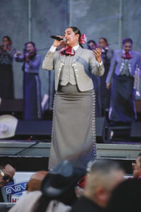 Female mariachi singer in a gray suit with silver embellishments and a red bow, singing into a microphone onstage with backup musicians in the background.
