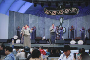 Mariachi group performs on a stage with microphones, gray jackets, and a large decorative treble clef backdrop; audience seated in front.