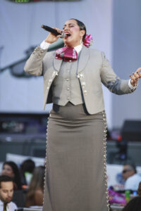 Female singer performing on stage, mic in hand, wearing a gray suit with pink bow and decorative silver buttons, mid-song.