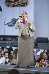 Female singer on a stage in a gray mariachi-inspired suit, pink bow, singing into a microphone with an audience watching.