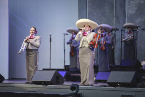 Mariachi band performing on stage in gray outfits; trumpet player on the left and violinist-singer center with a large sombrero, other musicians in background.