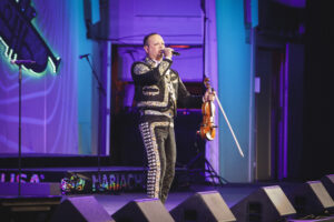 Male performer in an ornate black-silver mariachi-style suit sings into a handheld mic while holding a violin and bow on a purple-lit stage.
