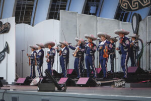 Mariachi musicians in blue outfits and large sombreros perform on a stage with violins and guitars in front of a decorative backdrop.
