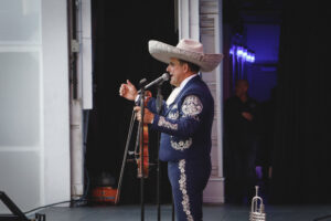 Mariachi performer in a blue embroidered suit and oversized sombrero sings into a microphone on stage with a guitar nearby.