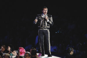 Male singer in a black and gold embroidered mariachi-style outfit performs on a lit stage, microphone in hand, audience in darkness beyond him