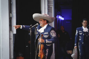 Mariachi performer in a blue embroidered suit and large sombrero, holding a violin and pointing to the audience on stage.