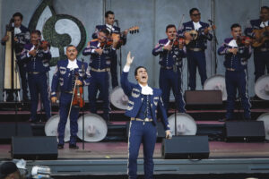 Male mariachi singer in a blue suit raises his hand while leading a band on a stage with violinists and guitar players in the background.