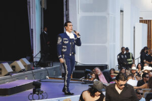 Male singer in a blue embroidered suit performs on a stage with a microphone, as a crowd watches from the audience area.