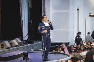 Male mariachi singer in a blue embroidered suit performs on stage outdoors for a crowd.