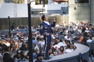 Male mariachi-style singer in a blue embroidered suit performs on an outdoor stage, microphone in hand, with a large crowd watching at a daytime show.