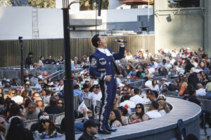 Male mariachi singer in a blue embroidered suit performs on a curved outdoor stage as a large audience watches from surrounding seating.