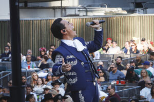 Male singer in a blue ornate suit performs on an outdoor stage with a microphone, engaging a large audience behind him.