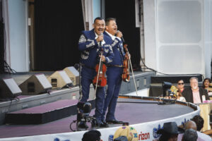 Two male mariachi performers in matching embroidered blue suits sing into microphones on a stage, each holding a violin, with an audience watching nearby.
