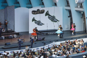 Mariachi band performs on an outdoor stage with dancers in bright dresses while an audience watches from seating below.
