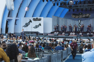 Audience watching a large band on an outdoor stage under blue arches; musicians play brass and percussion as a singer performs at a microphone in front of the crowd.